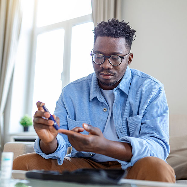 African man is sitting at the sofa and taking blood from his finger due to diabetes. The daily life of a man of African-American ethnicity person with a chronic illness who is using glucose tester.